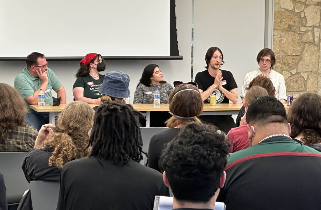 Five panelists sit at a table in front of a crowd. One is speaking while the others listen intently.