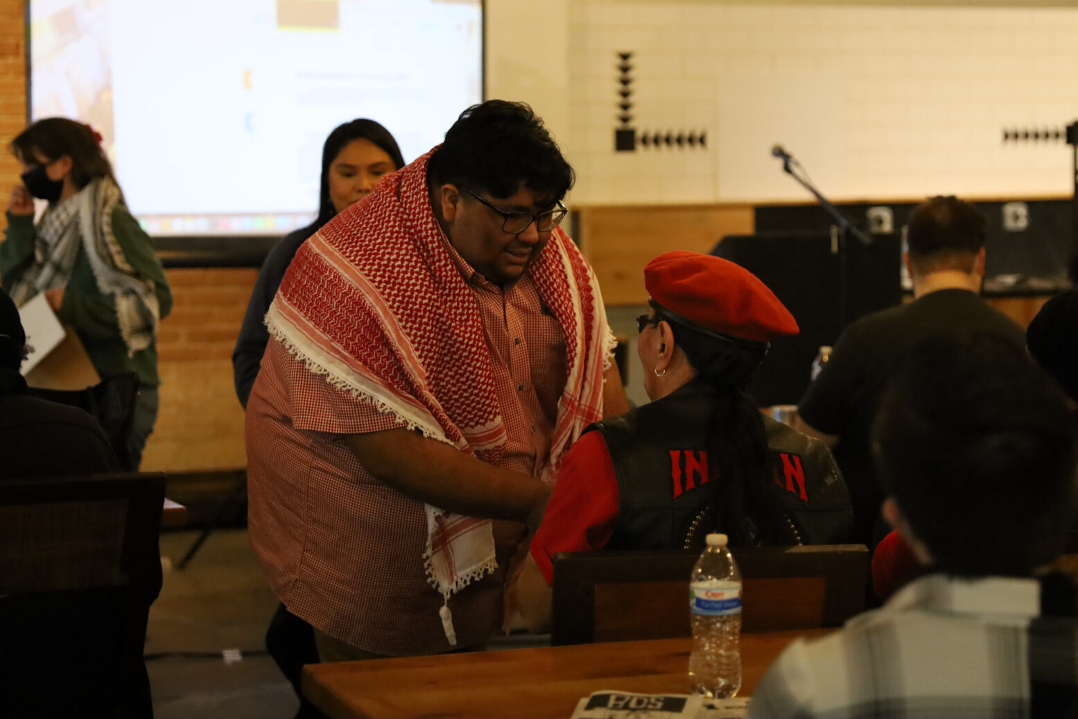 DSA member wearing a keffiyeh greets a member of the American Indian Movement who is wearing a red beret. A presentation screen and podium can be seen in the background.