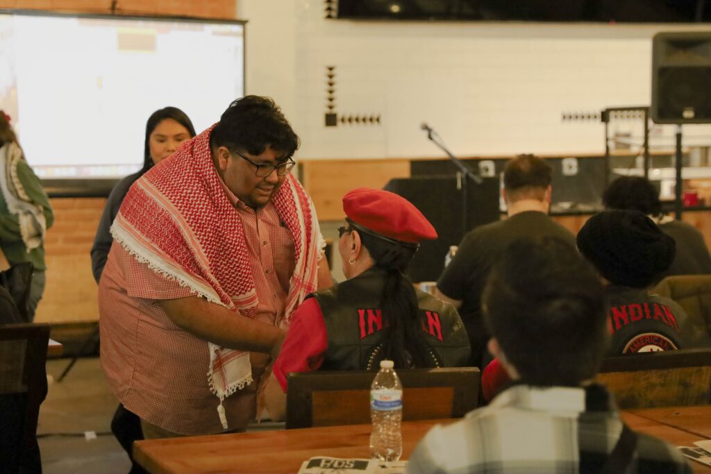 DSA member wearing a keffiyeh greets a member of the American Indian Movement who is wearing a red beret. A presentation screen and podium can be seen in the background.