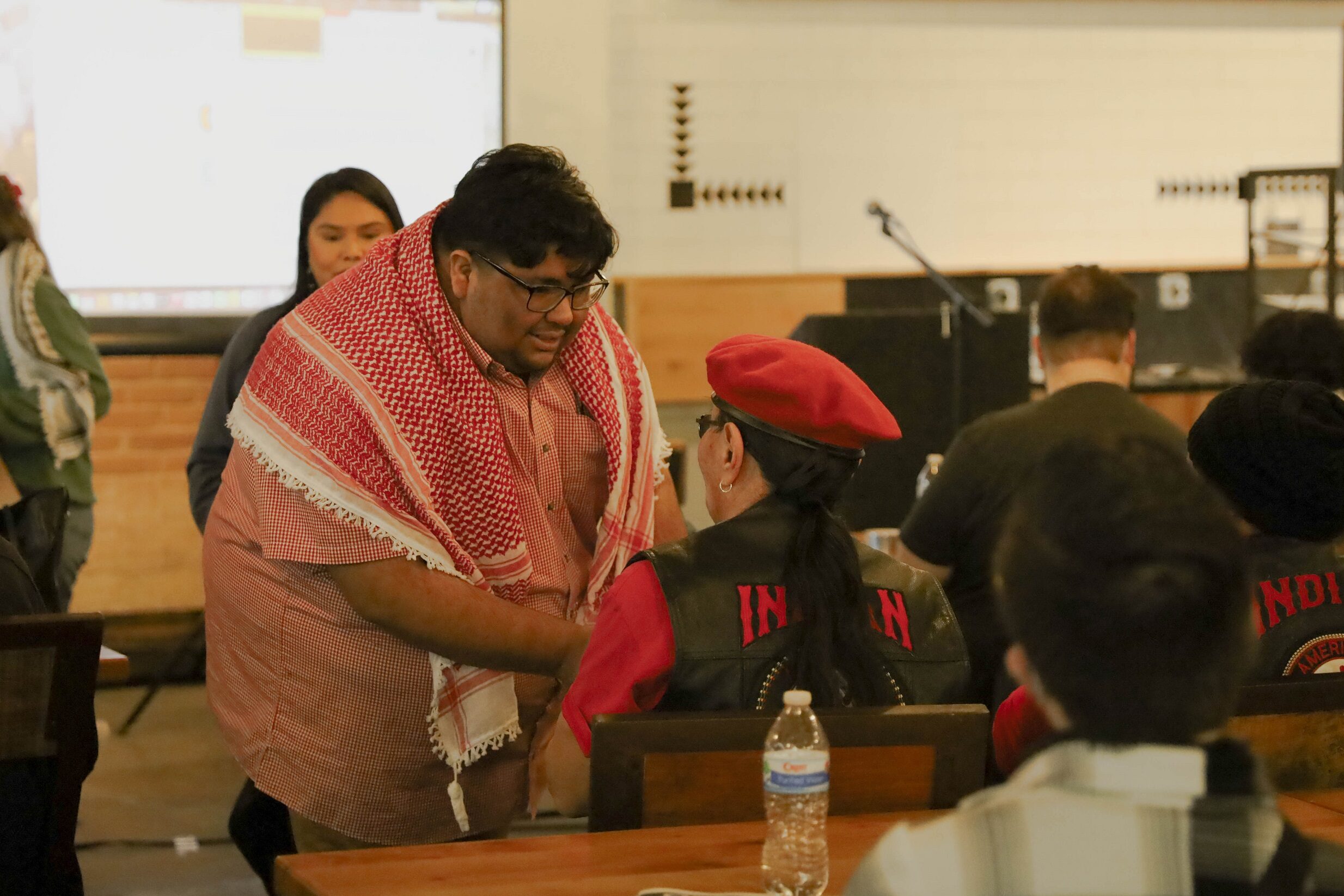 DSA member wearing a keffiyeh greets a member of the American Indian Movement who is wearing a red beret. A presentation screen and podium can be seen in the background.